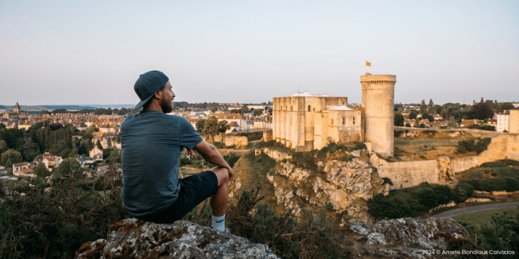 Wanderer sitzt an Klippe vor Panorama mit Burg in der Normandie in Frankreich